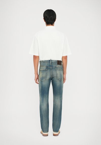 Jeans in denim chiaro con taglio dritto, indossati in vita, dotati di una toppa in pelle marrone e con leggeri sbiadimenti. Camicia bianca a maniche corte.