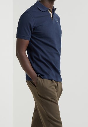 Mężczyzna w granatowej koszulce polo z krótkim rękawem i detalem w kratę na kołnierzyku oraz w brązowych spodniach, stojący z jedną ręką w kieszeni na białym tle.