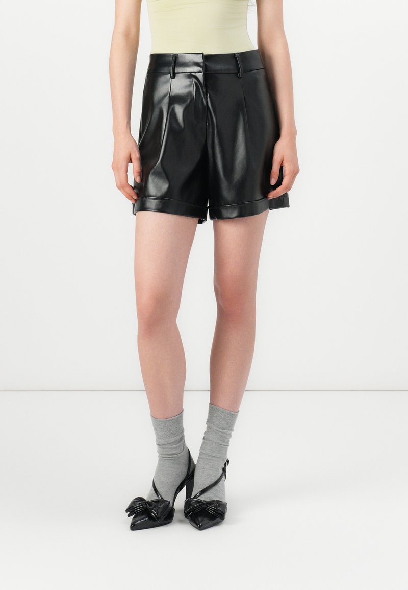 Svart lærshorts med plissert front, rullet kant og en jevn tekstur. Kombinert med grå sokker og svarte spisse hæler med sløyfer.