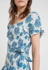 Blå och grön blommig mönsterklänning med korta puffärmar, fyrkantig halsringning och en knytning i midjan. Tillverkad av lättviktig tyg.