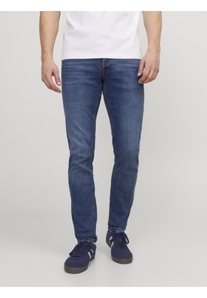 Homme portant un jean bleu slim, des baskets navy avec des rayures bleu clair, et une chemise blanche, se tenant devant un fond gris clair uni.