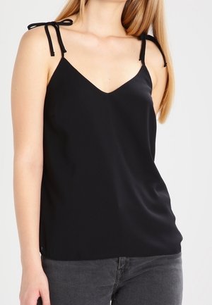 Woman wearing a sleeveless black camisole top with thin shoulder ties and dark gray jeans against a plain background.