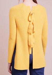 Gelber gerippter Strickpullover mit langen Ärmeln, offenes Rückendesign mit Schleifenakzenten und geschlitztem Saum für zusätzliche Details.