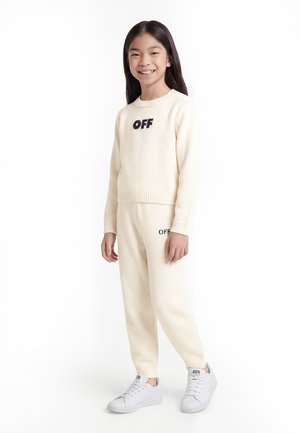 Šypsanti mergina su ilgais juodais plaukais, dėvinti kreminės spalvos megztinį ir kelnių komplektą su užrašu „OFF“, dėvinti baltus sportbačius, stovinti ant balto fono.