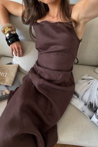 Ruskea pellava-mekko, jossa on löysä istuvuus, spagettiolkaimet ja solmittava yksityiskohta. Mekossa on teksturoitu kangas ja kerroksittain laskeutuvia yksityiskohtia. Mukana on kuviollisia asusteita.