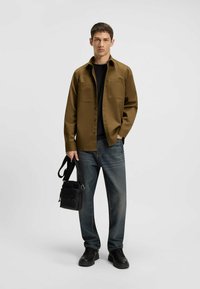 Bruin button-up shirt met twee voorzakken, gedragen over een zwart T-shirt. Blauwe jeans met vervaging, zwarte sneakers en een zwarte schoudertas.