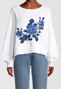 Sudadera blanca de manga larga con rosas bordadas en azul en la parte frontal, combinada con unos vaqueros de tiro alto en azul claro.