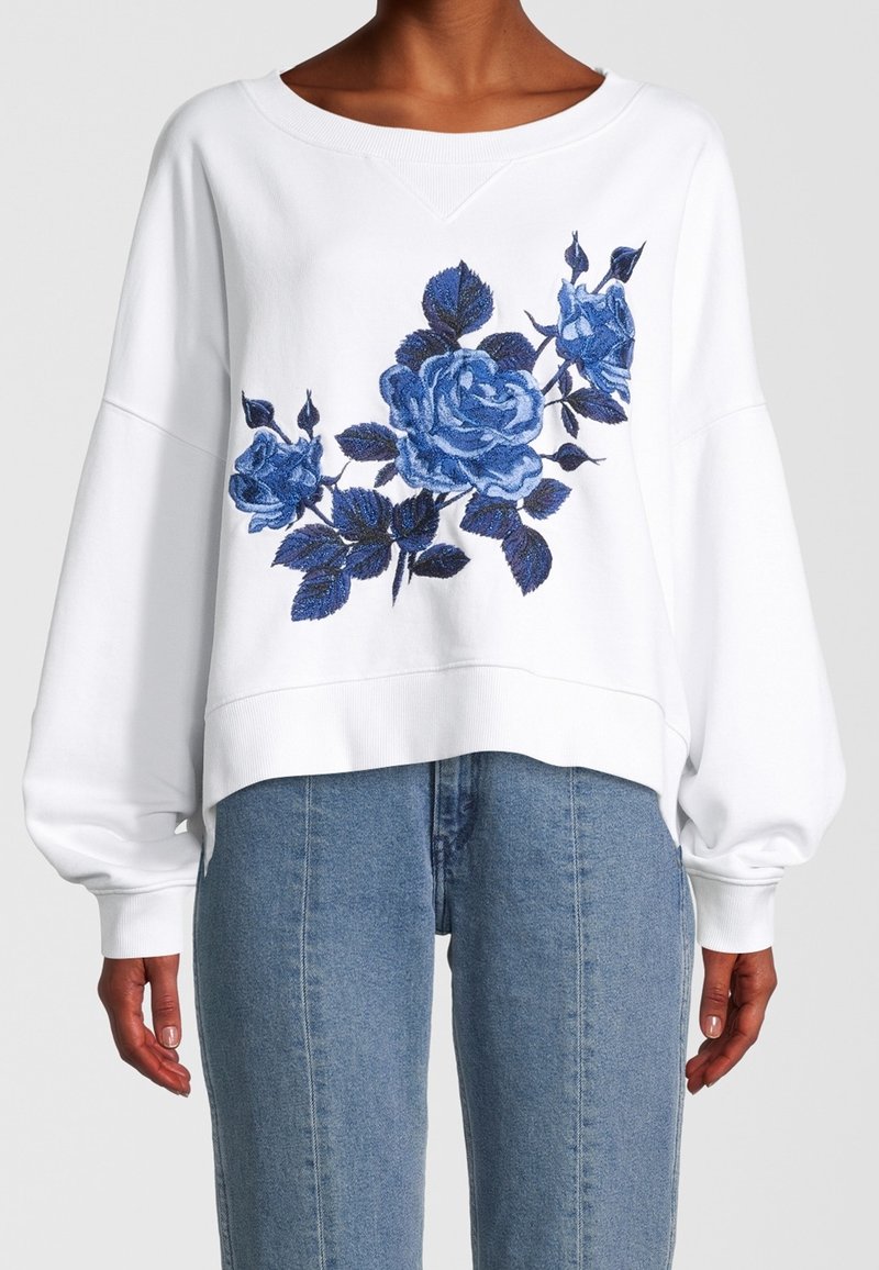 Sudadera blanca de manga larga con rosas bordadas en azul en la parte frontal, combinada con unos vaqueros de tiro alto en azul claro.