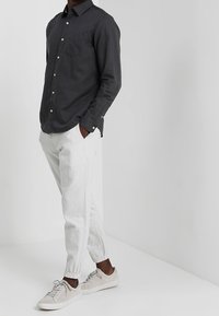 Man in een zwart lange mouwen overhemd met knopen, witte jogger pants en lichtbeige sneakers, staande met handen in de zakken tegen een witte achtergrond.