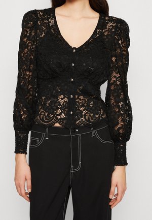 Frau trägt eine schwarze Spitzenbluse mit langen Ärmeln und Knöpfen sowie eine hoch taillierte schwarze Hose mit weißen Nähten.