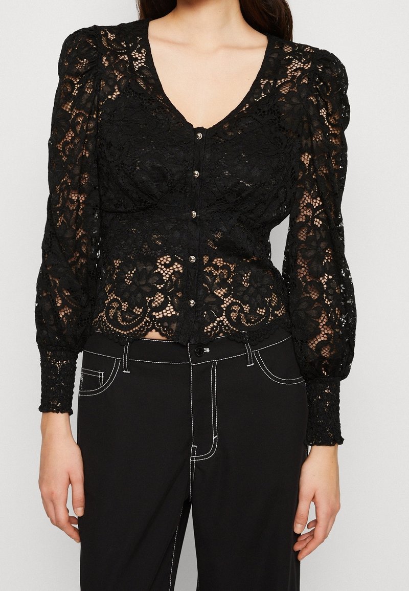 Femme portant un chemisier noir en dentelle à manches longues avec des boutons et un pantalon noir taille haute avec des coutures blanches.