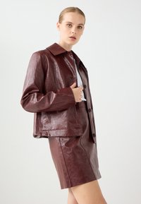 Bordeaux nep leren jasje met een kraag, knoopsluiting en een relaxte pasvorm. Gelaagde oppervlakte met een bijpassende korte rok.