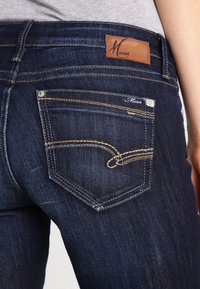 Donkerblauwe denim jeans met een kenmerkend leren label, koperen stiksels en een uniek zakontwerp met een gebogen naaddetail.