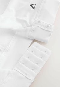 Vêtements de sport en tissu blanc avec des bretelles réglables à agrafes et un logo triangulaire près de la couture sur un matériau lisse et en maille.