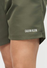 Nærbilde av en person som har på seg olivengrønne shorts med hvit «Calvin Klein»-logo over kneet på høyre ben.