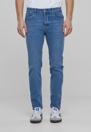 Kék farmernadrág, slim fit szabással, öt zsebbel, gombos záródással és finom kopással a lábak mentén. Sportcipővel viselve.