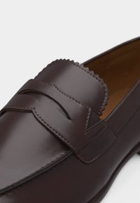 Mocassin en cuir brun foncé avec détail de bord ondulé et une lanière horizontale sur le coup-de-pied, présenté sur un fond clair.