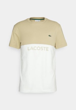Kortärmad t-shirt i beige och vitt, med en grön krokodil-logotyp och "LACOSTE" tryckt i beige över framsidan.