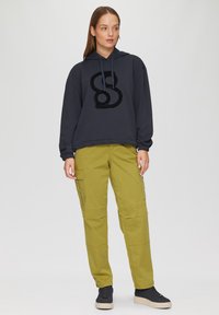 Sweat à capuche oversized gris foncé avec un logo noir texturé, associé à un pantalon cargo vert olive fuselé et des baskets noires à semelles crème.