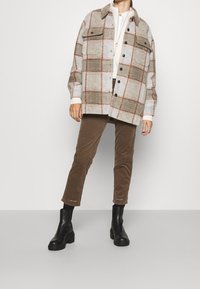 Chemise flanelle à carreaux dans des tons gris, marron et crème atténués avec une coupe décontractée, associée à un pantalon en velours côtelé marron et des bottines noires.