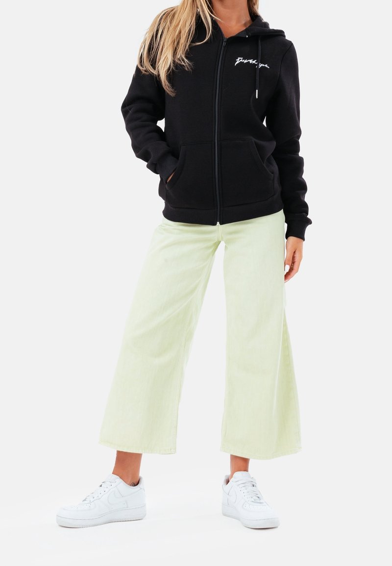 Femme portant un sweat à capuche noir zippé avec des poches avant, un pantalon droit clair et large, et des baskets blanches, debout devant un fond uni.