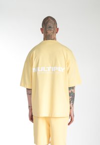 Kort gul oversized t-shirt med rund hals og hvid "MULTIPLY" logo på ryggen; bæres med matchende shorts.