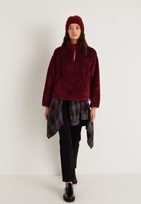 Bordowy polar z półzamkiem, zestawiony z czarnymi spodniami i koszulą w kratę przewiązaną wokół talii. Model nosi pasującą czapkę beanie.