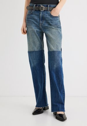 Kobieta ubrana w dwukolorowe, proste niebieskie dżinsy, czarny pasek z ćwiekami, czarną bluzkę i czarne buty na obcasie, stojąca z jedną ręką w kieszeni.