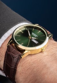 Montre dorée avec un cadran vert, un bracelet en cuir marron et un affichage de la date. Elle possède des index des heures fins et un bracelet texturé. Résistante à l'eau jusqu'à 50 mètres.