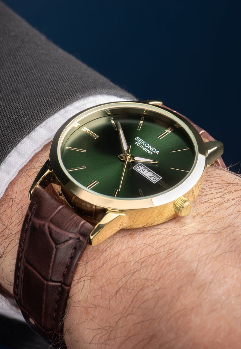 Montre dorée avec un cadran vert, un bracelet en cuir marron et un affichage de la date. Elle possède des index des heures fins et un bracelet texturé. Résistante à l'eau jusqu'à 50 mètres.