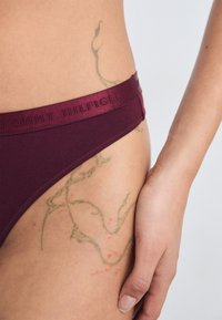 Burgundy cotton hipster underwear with a textured logo waistband. Features visible skin and a green tattoo on the hip area.