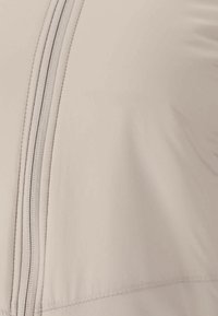 Lys beige, blødt stof med en central lynlås og glat tekstur. Designet er tætsiddende med rene syninger.