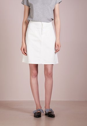 Persona con camiseta gris de manga corta, falda blanca hasta la rodilla y zapatos planos negros con detalles de lazo de cuadros, de pie contra un fondo pálido.