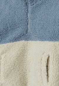 Fleece-Pullover mit Reißverschluss am Hals, hellblauem oberen Teil und cremefarbenem unteren Teil, mit einer strukturierten, weichen Oberfläche und seitlichen Taschen.