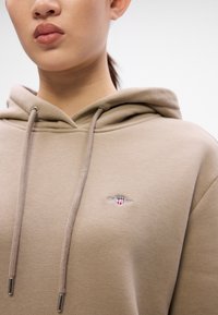 Bež hoodie s vezicama, prednjim džepom i malim veznim logom na prsima. Mekana, teksturirana tkanina s opuštenim krojem.