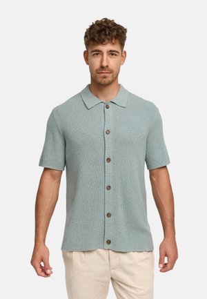 Man draagt een lichtblauw gebreid overhemd met korte mouwen en knopen en een beige broek, staand tegen een effen witte achtergrond.