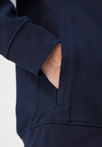 Main reposant à l'intérieur de la poche avant d'un sweat-shirt bleu marine avec un bord-côtes au niveau de la manche.