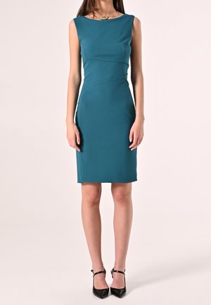 Kvinde iført ærmeløs teal knælængde sheath-kjole med asymmetrisk søm og sorte ankelremssandaler, stående mod en ensfarvet baggrund.