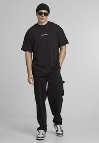Sort oversized T-shirt med teksten "Another Cotton", parret med tapered sorte cargo bukser med sidelommer og en afslappet pasform. Sort beanie og solbriller fuldender looket.