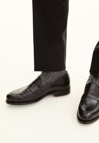 Schwarze Leder-Schluppen mit glatter Oberfläche, ausgestattet mit einer erhöhten Naht und niedrigem Absatz, kombiniert mit grau gemusterten Socken. Die Hose ist schwarz.