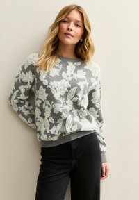 Suéter gris con un patrón floral esponjoso en blanco y gris claro. Cuello redondo, mangas largas, dobladillo acanalado. Combinado con jeans negros.