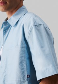 Camicia azzurro chiaro a maniche corte con colletto appuntito, taschino sul petto e piccola toppa con logo vicino all'orlo. Tessuto di cotone liscio.