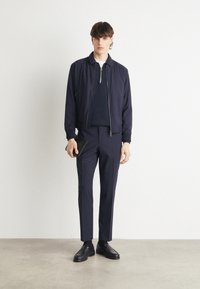 Mornarska zip-up bomber jakna preko mornarskega puloverja, v kombinaciji z oprijetimi mornarskimi hlačami in črnimi usnjenimi čevlji. Model drži črno torbico.