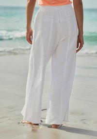 Pantalones anchos de lino blanco con un ajuste holgado, que cuentan con una cinturilla elástica y bolsillos mínimos, de pie sobre arena mojada junto al océano.