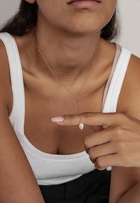 Złoty naszyjnik z jednym białym perłowym wisiorkiem, noszony na nagiej szyi, na jasnoszarym tle, z ręką trzymającą wisiorek.