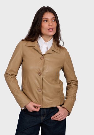 Žena s dlouhými tmavými vlasy, oblékající hnědou koženou bundu na knoflíky přes bílou košili s límečkem a tmavě modré džíny, pózující s rukama u kapes.