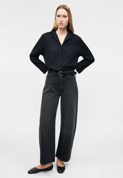 Γυναίκα όρθια με τα χέρια στη μέση, φοράει μαύρο πουκάμισο με κουμπιά, ψηλόμεσο παντελόνι με φαρδιά μπατζάκια σε σκούρο χρώμα, μαύρη ζώνη και μαύρες μπαλαρίνες.