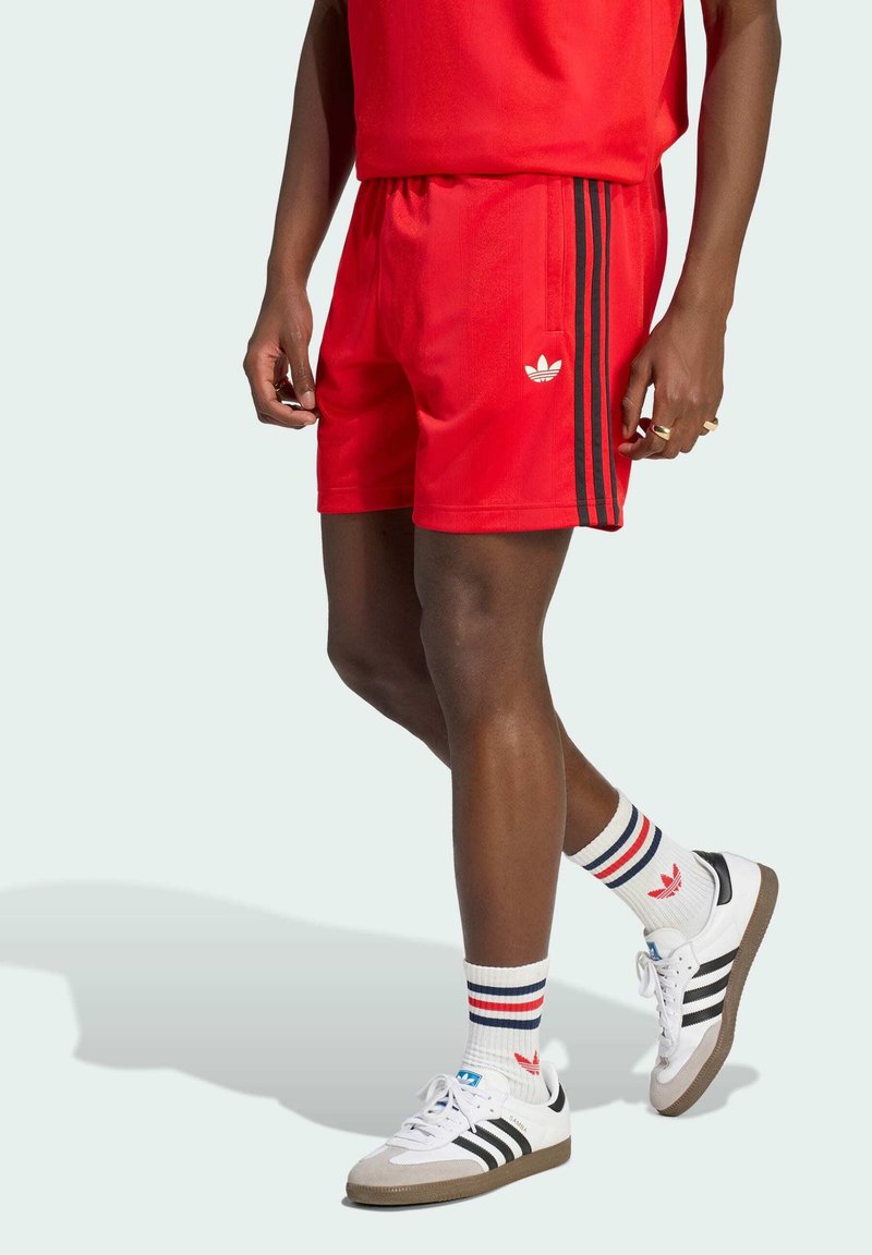 Muž nosí červené kraťasy Adidas s černými pruhy, bílé ponožky Adidas s červenými a modrými pruhy a bílé tenisky Adidas s černými pruhy.