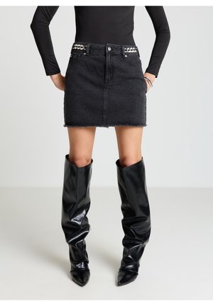Frau trägt ein schwarzes langärmliges Oberteil, einen schwarzen Denim-Minirock mit Kettenakzenten und glänzende schwarze kniehohe Stiefel.