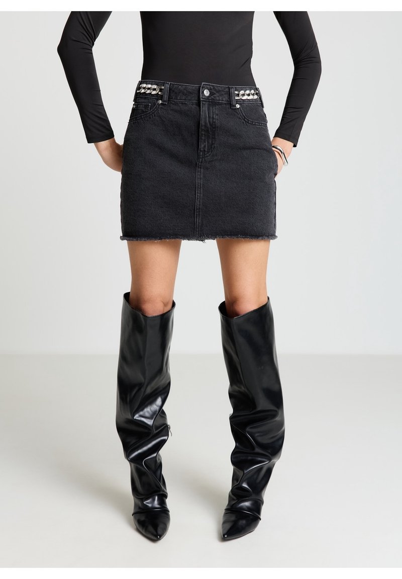 Femme portant un haut noir à manches longues, une mini-jupe en jean noire avec des accents de chaînes et des bottes noires brillantes montant jusqu'aux genoux.